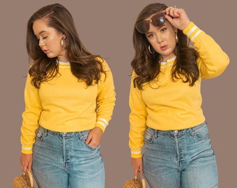 May include: Two photographs of a person wearing a yellow long-sleeved sweater with white stripes on the cuffs and collar. The person is wearing blue jeans and has long brown hair. In one photo, the person is looking down, and in the other photo, the person is wearing sunglasses and looking to the side.