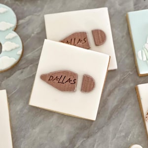 May include: Two white square cookies decorated with brown fondant footprints. The top cookie has the word "DALLAS" written on the footprint. The bottom cookie has the word "MILLAS" written on the footprint.