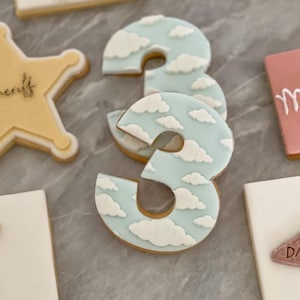 May include: Two blue and white cookies decorated with a cloud pattern, shaped like the number three. The cookies are stacked on top of each other.