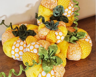 May include: A collection of handmade fabric pumpkins in various sizes. The pumpkins are primarily yellow with white polka dots and orange floral patterns. Green fabric leaves and curly green ribbon add detail.