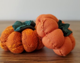May include: Two small, handmade orange fabric pumpkins with green fabric stems. The pumpkins have a textured appearance and are likely used for fall decor. The pumpkins are sitting on a wooden surface.