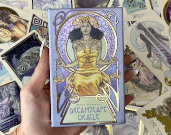 May include: A blue and gold oracle deck box with the title "Dreamscape Oracle" and the name "Matt Hughes" printed on the front. The box features an illustration of a woman with dark hair and a gold crown, sitting on a throne with scales in her hands.
