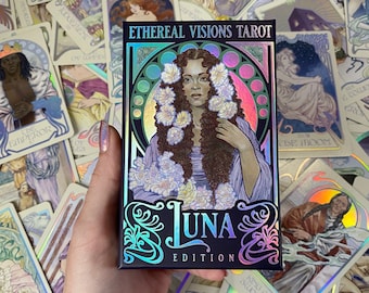 May include: A rectangular tarot deck box titled Ethereal Visions Tarot Luna Edition. The box features a holographic illustration of a person with long dark hair adorned with white flowers, set against a circular frame with ornate, swirling borders.