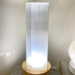 Puede incluir: Una lámpara rectangular de selenita blanca translúcida con un interior brillante. La lámpara se asienta sobre una base redonda de madera clara. La lámpara mide aproximadamente 20 cm de altura.