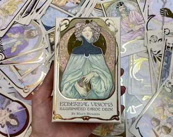 May include: A white box with gold accents featuring a tarot deck called "Ethereal Visions Illuminated Tarot Deck" by Matt Hughes. The box shows a woman with dark hair and a blue dress holding a golden cup.