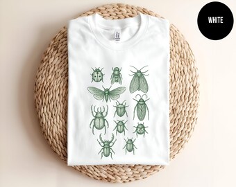 May include: A white t-shirt featuring a central graphic print of various insects in a dark green ink. The illustration includes detailed drawings of beetles, a moth, and other bugs arranged in a cluster on the front of the garment.
