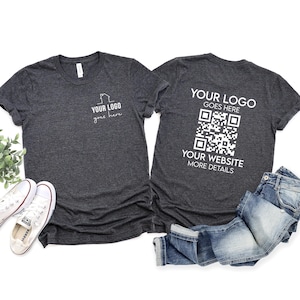 May include: Two dark heather gray t-shirts featuring white custom printing. The left shirt displays a small house icon with the text YOUR LOGO goes here. The right shirt features the text YOUR LOGO GOES HERE, a QR code, and YOUR WEBSITE MORE DETAILS.