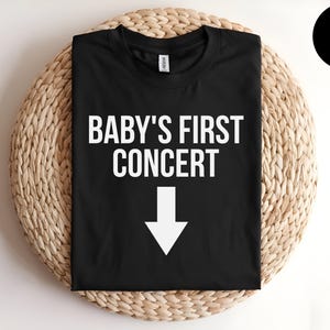 Pode incluir: Camiseta preta com texto branco que diz "BABY'S FIRST CONCERT" com uma seta branca apontando para baixo.
