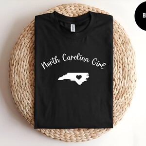 Pu&ograve; includere: Maglietta nera con la scritta "North Carolina Girl" in bianco. Un contorno bianco della Carolina del Nord con un cuore &egrave; centrato sotto il testo. La maglietta &egrave; esposta su una superficie rotonda e intrecciata.