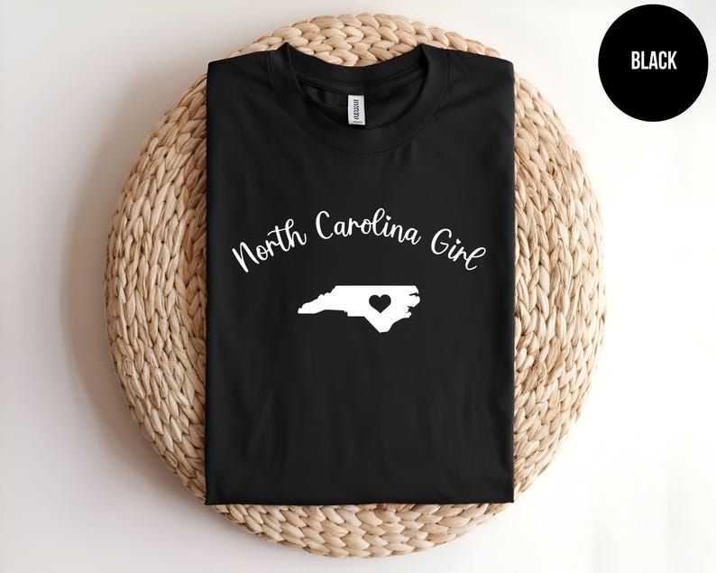 Pu&ograve; includere: Maglietta nera con la scritta "North Carolina Girl" in bianco. Un contorno bianco della Carolina del Nord con un cuore &egrave; centrato sotto il testo. La maglietta &egrave; esposta su una superficie rotonda e intrecciata.
