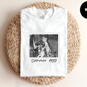 May include: White t-shirt featuring a black and white image of a child on a toy horse, with the text "Summer 1980" printed below the image. The shirt is displayed on a woven, circular surface.