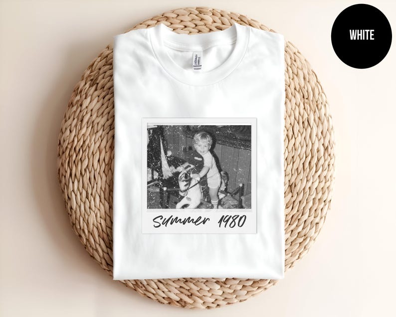 May include: White t-shirt featuring a black and white image of a child on a toy horse, with the text "Summer 1980" printed below the image. The shirt is displayed on a woven, circular surface.