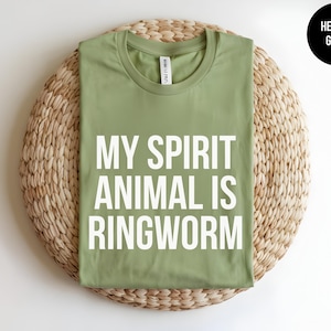 May include: A heather green crew neck t-shirt featuring the white text MY SPIRIT ANIMAL IS RINGWORM printed in a bold, sans-serif font on the front. The shirt has a standard short-sleeve design and a visible brand label at the inner neckline.