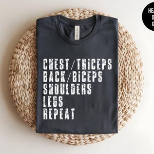 May include: A heather dark grey short-sleeve t-shirt featuring white distressed text that reads: CHEST/TRICEPS, BACK/BICEPS, SHOULDERS, LEGS, REPEAT. The shirt has a crew neckline and a standard fit, with a label visible at the inner collar.