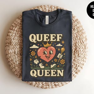 May include: Heather dark grey t-shirt with a retro graphic. The design features a smiling heart wearing a crown, holding a scepter, and the words "QUEEF QUEEN" in a vintage font. The shirt is folded on a woven mat.