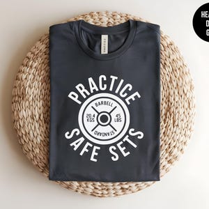 May include: A dark heather grey t-shirt with a white graphic of a weight plate that says "Practice Safe Sets" with the words "Barbell" and "Standard" on the weight plate.