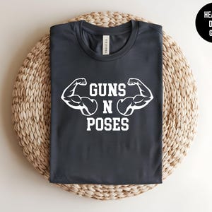 May include: A heather dark grey t-shirt with white text that reads "Guns N Poses" and two white bicep illustrations.