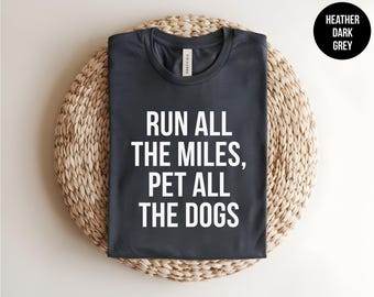 May include: A heather dark grey crew neck t-shirt featuring the white text RUN ALL THE MILES, PET ALL THE DOGS printed in a bold sans-serif font on the front. The shirt has short sleeves and a standard fit, with a label visible at the neckline.