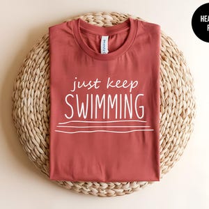 May include: A heather red t-shirt with the text "just keep swimming" in white lettering. The shirt is on a woven straw surface.