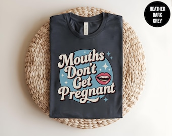 May include: A heather dark grey t-shirt featuring a light blue circular graphic with white star accents and a pink lip illustration. The text "Mouths Don't Get Pregnant" is printed in a bold, white, retro-style font across the center of the design.