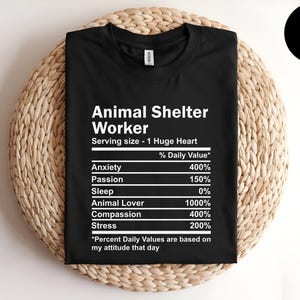 May include: Black t-shirt with white text that reads "Animal Shelter Worker" and a nutrition label listing "Anxiety", "Passion", "Sleep", "Animal Lover", "Compassion", and "Stress" with percentages next to each item.
