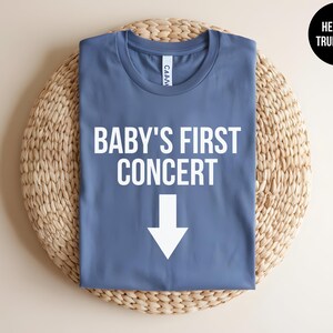 Pode incluir: Uma camiseta azul com o texto "BABY'S FIRST CONCERT" e uma seta branca apontando para baixo.