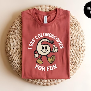 May include: A heather red crew neck t-shirt featuring a graphic of a cartoon character shaped like a colonoscope with a smiling face, giving a thumbs up, and wearing boots. The text reads I GET COLONOSCOPIES FOR FUN in white capital letters.