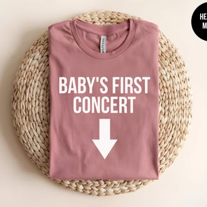Pode incluir: Uma camiseta cor malva com o texto "Baby's First Concert" e uma seta branca apontando para baixo.