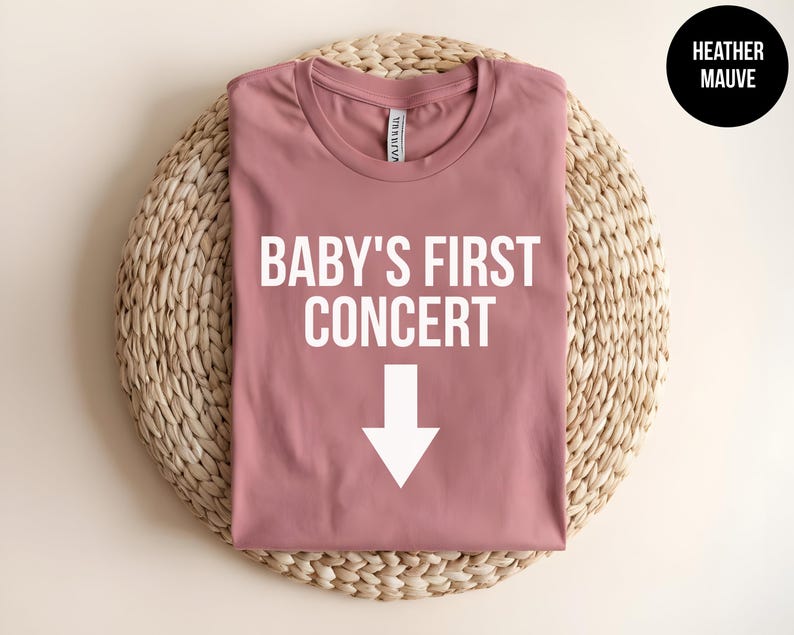 Pode incluir: Uma camiseta cor malva com o texto "Baby's First Concert" e uma seta branca apontando para baixo.