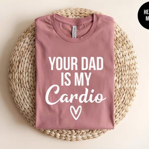 May include: A heather mauve short-sleeve t-shirt with white text that reads "YOUR DAD IS MY Cardio" above a small white heart outline. The shirt features a crew neckline and is displayed folded, with a circular black tag in the corner.