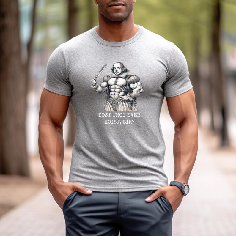 Op de afbeelding: Een grijs T-shirt met een zwart-witte illustratie van een gespierde William Shakespeare die een pen vasthoudt. De tekst "Dost thou even hoist, sir?" staat onder de illustratie.