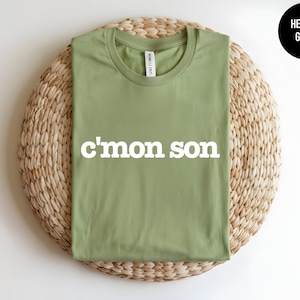 May include: A heather green crew neck t-shirt featuring the white text c'mon son printed across the chest. The shirt has short sleeves and a standard fit, with a small white brand label visible inside the collar at the neckline.