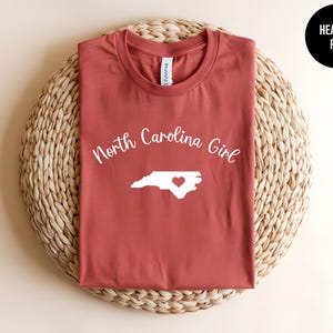 Pu&ograve; includere: T-shirt rosso melange con la scritta "North Carolina Girl" sopra un contorno bianco dello stato con un cuore. La maglietta &egrave; esposta su un tappetino circolare intrecciato. La maglietta &egrave; rossa.