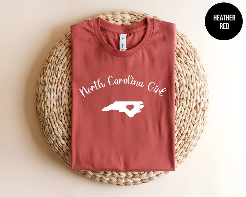 Pu&ograve; includere: T-shirt rosso melange con la scritta "North Carolina Girl" sopra un contorno bianco dello stato con un cuore. La maglietta &egrave; esposta su un tappetino circolare intrecciato. La maglietta &egrave; rossa.