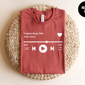 May include: A heather red t-shirt featuring a white graphic of a music player interface. The design includes the text Custom Song Title and Artist Name, a heart icon, a progress bar, and playback controls including shuffle, skip, play, and repeat.