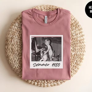 May include: Heather mauve t-shirt with a black and white photo print. The photo shows a child on a rocking horse with the text "Summer 1980". The shirt is displayed on a woven, circular mat.