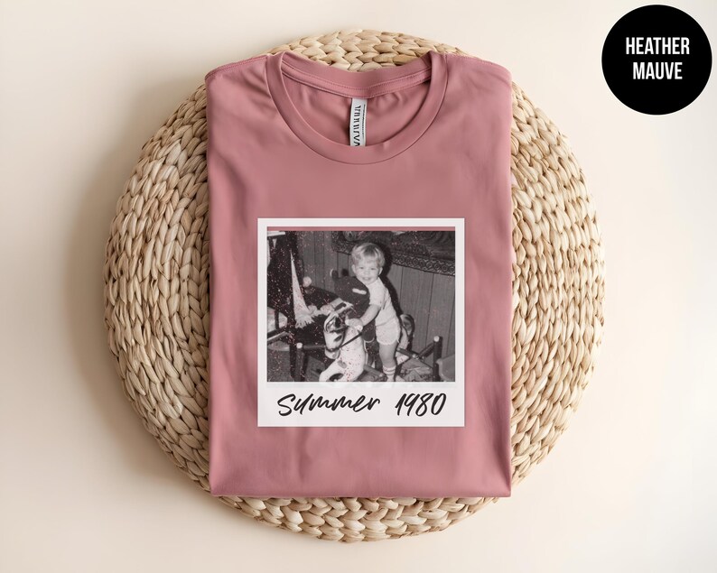 May include: Heather mauve t-shirt with a black and white photo print. The photo shows a child on a rocking horse with the text "Summer 1980". The shirt is displayed on a woven, circular mat.