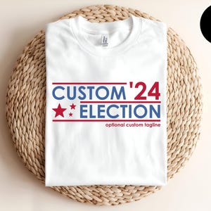 May include: A white t-shirt featuring the blue and red text CUSTOM '24 ELECTION with three red stars on the left side. Below the main text, the phrase optional custom tagline is printed in red. The design is centered on the front of the shirt.