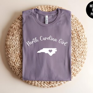 Pu&ograve; includere: Una maglietta viola erica con la scritta "North Carolina Girl" e un contorno bianco dello stato con un cuore. La maglietta &egrave; esposta su un sottobicchiere circolare intrecciato. Un cerchio nero con testo bianco recita "Heather Team Purple".