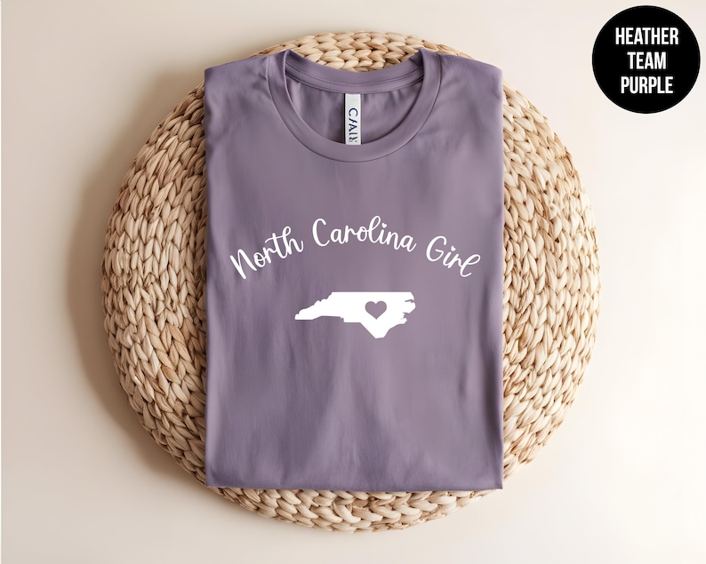 Pu&ograve; includere: Una maglietta viola erica con la scritta "North Carolina Girl" e un contorno bianco dello stato con un cuore. La maglietta &egrave; esposta su un sottobicchiere circolare intrecciato. Un cerchio nero con testo bianco recita "Heather Team Purple".