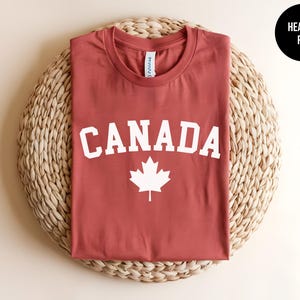 May include: A heather red crew neck t-shirt featuring the word CANADA in white capital letters arched above a white maple leaf graphic. The shirt has a white inner neck label and is displayed flat, showcasing the front design clearly.
