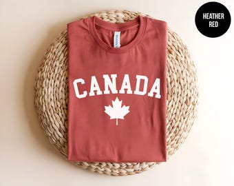 May include: A heather red crew neck t-shirt featuring the word CANADA in white capital letters arched above a white maple leaf graphic. The shirt has a white inner neck label and is displayed flat, showcasing the front design clearly.