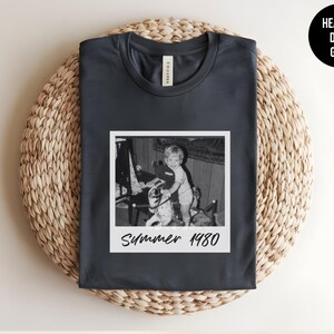 May include: Heather dark grey t-shirt with a vintage black and white photo print. The photo is a Polaroid-style image of a child with a toy, with the text "Summer 1980" below the image. The shirt is laid on a woven mat.