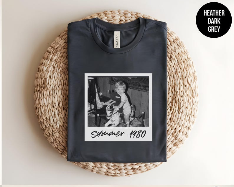 May include: Heather dark grey t-shirt with a vintage black and white photo print. The photo is a Polaroid-style image of a child with a toy, with the text "Summer 1980" below the image. The shirt is laid on a woven mat.
