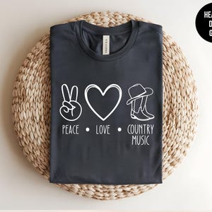 May include: Heather dark grey t-shirt with a white peace sign, heart, cowboy hat and boots graphic and the text "Peace. Love. Country Music."