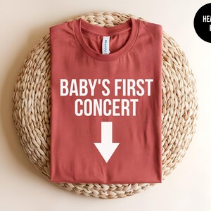 Pode incluir: Uma camiseta de cor vermelha heather com o texto "BABY'S FIRST CONCERT" e uma seta branca apontando para baixo.