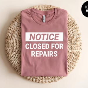 May include: A heather mauve t-shirt with the text "NOTICE CLOSED FOR REPAIRS" in white.