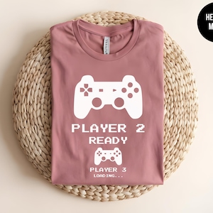 May include: A heather mauve short-sleeve t-shirt featuring a white graphic of a video game controller above the text PLAYER 2 READY, followed by a smaller controller icon and the text PLAYER 3 LOADING... in a pixelated white font style.