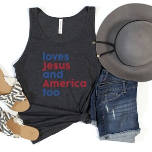 May include: A dark gray tank top with the words "loves Jesus and America too" in blue and red. The image also includes denim shorts, a gray hat, and striped sandals.