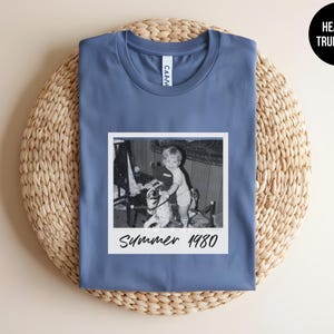 May include: A blue t-shirt with a vintage black and white photo of a child on a toy horse. The photo is in a white frame and has the text "Summer 1980" in a handwritten style. The shirt is folded on a woven mat.